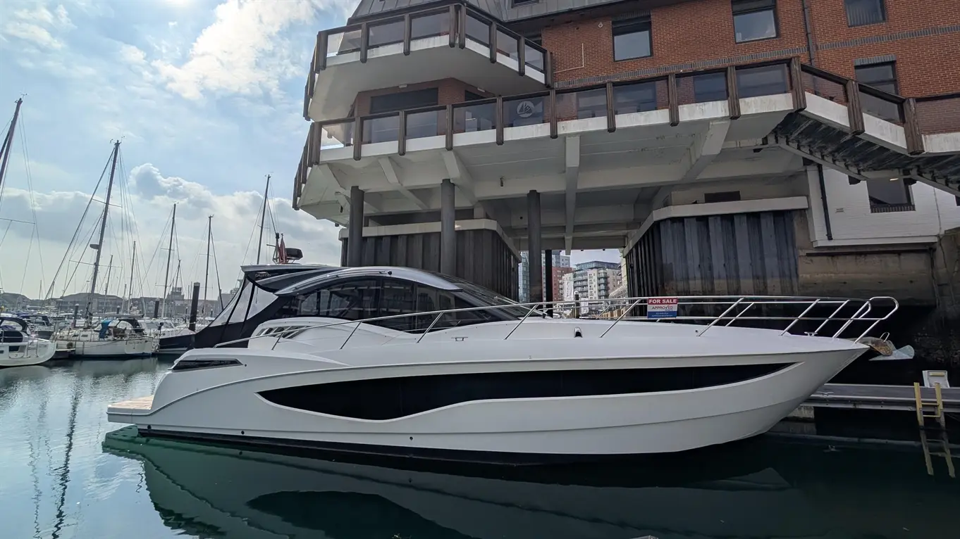 Luxury white motor yacht docked at a marina with tall sailboats and a modern building above, featuring a "For Sale" sign.